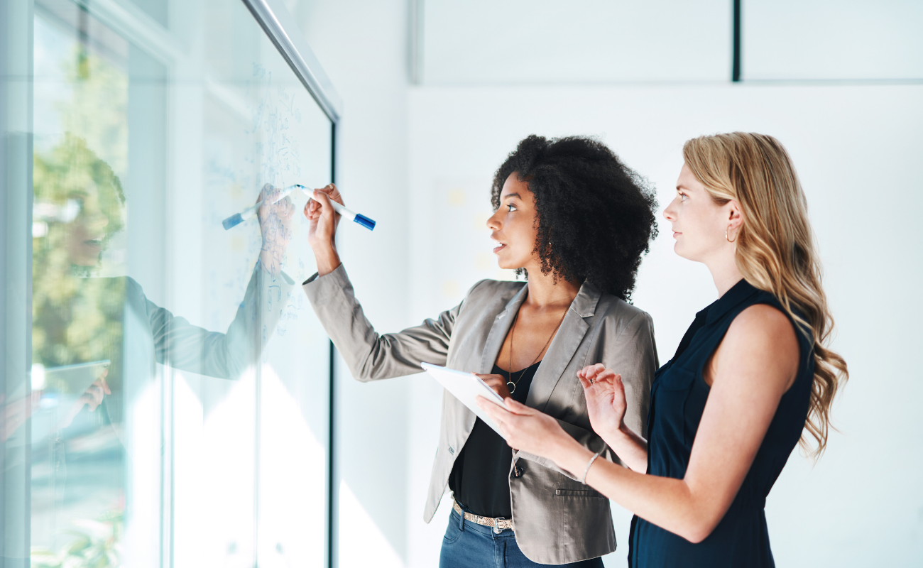 Two women collaborating on a glass whiteboard with an tablet and marker.