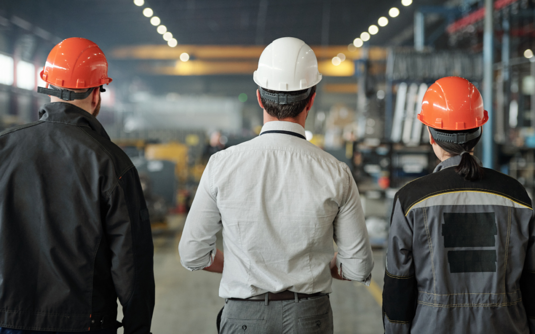Two factory workers and an executive with hard hats on overlooking a factory floor.