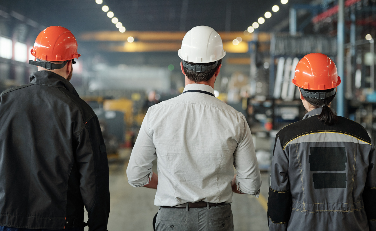 Two factory workers and an executive with hard hats on overlooking a factory floor.