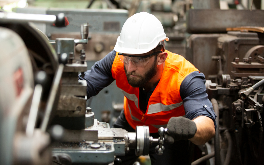 Manufacturer working on machine with hard hat, orange vest, gloves, and safety glasses.