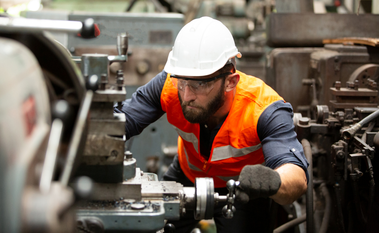 Manufacturer working on machine with hard hat, orange vest, gloves, and safety glasses.