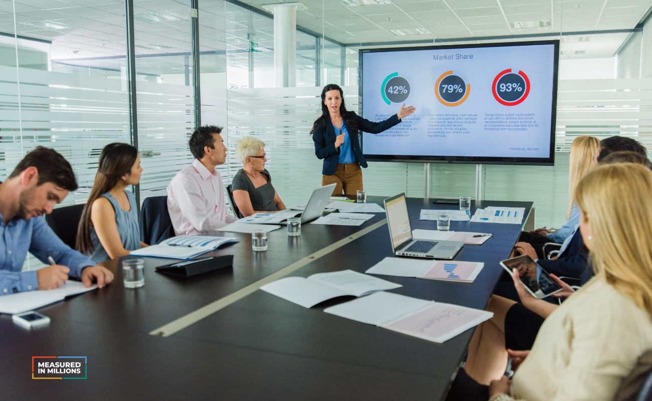 Woman presenting a slide to a room full of people at a conference table.